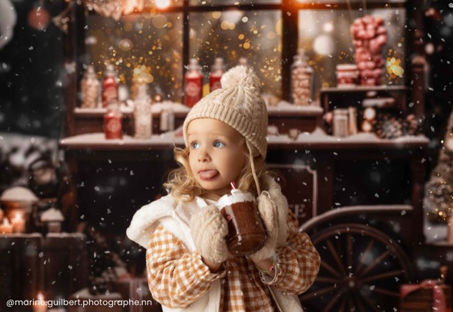 Enfant en bonnet et gants, marché de Noël en arrière-plan, neige artificielle, ambiance festive, gobelet de chocolat chaud, lumières de Noël